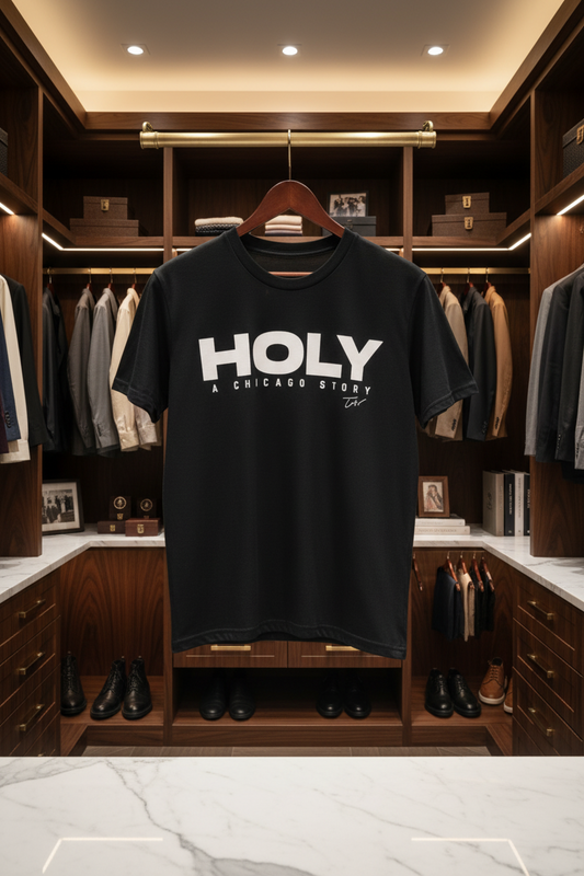 A black short-sleeve T-shirt hanging on a wooden hanger in a luxury walk-in closet. The shirt features the text 'HOLY A CHICAGO STORY' printed in bold white capital letters across the chest.